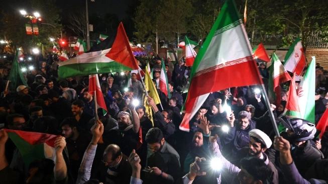 Demonstran mengibarkan bendera Iran dan bendera Palestina saat mereka berkumpul setelah Iran melancarkan serangan drone dan rudal ke Israel di depan Kedutaan Besar Inggris di Teheran, Iran, Minggu (14/4/2024) (sumber: suara.com)