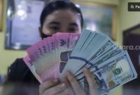 Ilustrasi petugas menunjukkan mata uang Rupiah dan Dolar AS di tempat penukaran uang. [Suara.com/Alfian Winanto]